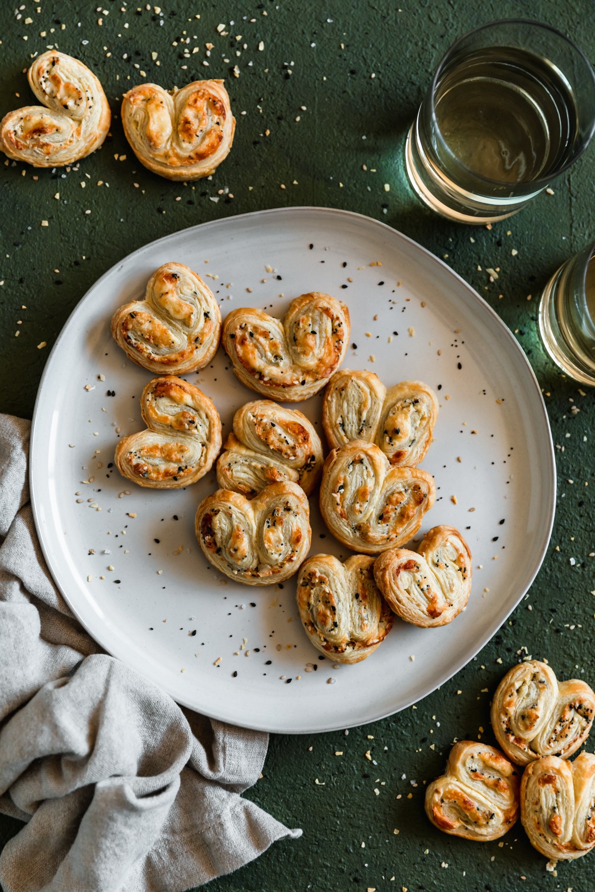 An overhead closeup image of a white plate of savory palmiers with parmesan and everything bagel spice on an emerald green table sprinkled with everything spice. In the right upper corner is two glasses of white wine. The plate is on a beige linen and there are more palmiers in the upper left and bottom right corners.