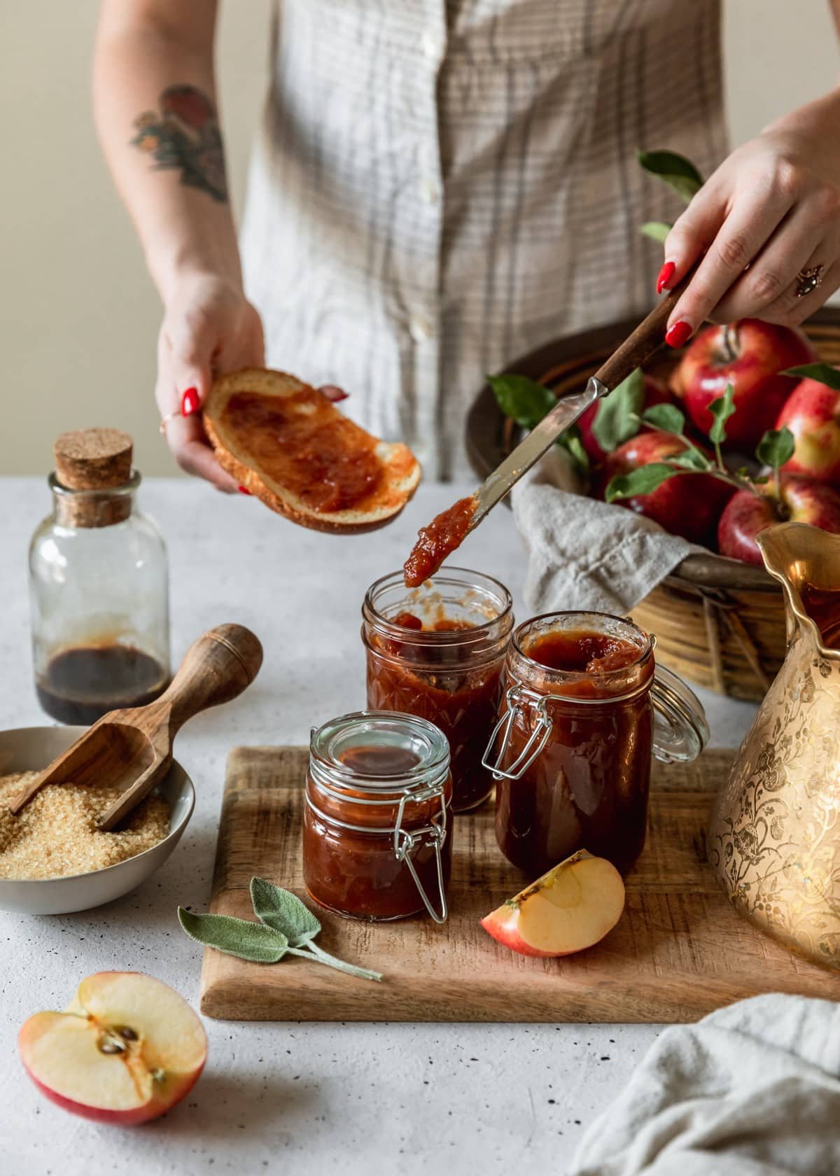 A side image of three jars of apple butter on a wood board next to a gold teapot. In the background is a basket of apples and a woman wearing a white and grey dress spreading apple butter on a slice of toast.