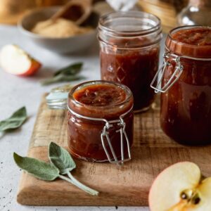 A side image of a small jar of apple butter on a wood board with two more jars in the background. the board is on a white counter with sliced apples, sage leaves, and a bowl of sugar in the background.