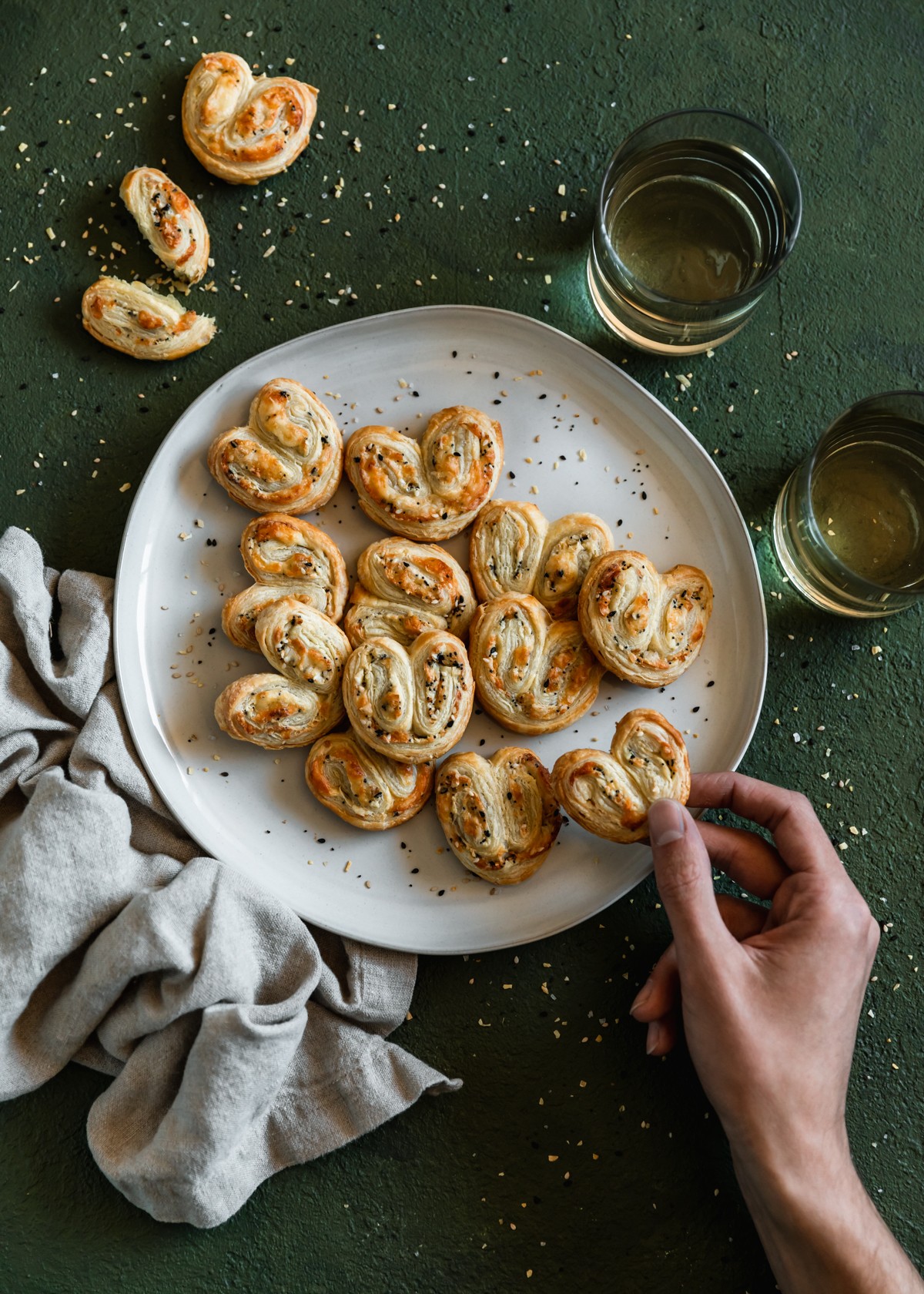 On overhead image of a man's hand grabbing a savory palmier with parmesan and everything spice from a white plate of palmiers. The plate is on a dark green table spinkled with everything bagel spice next to two glasses of white wine and a beige linen.