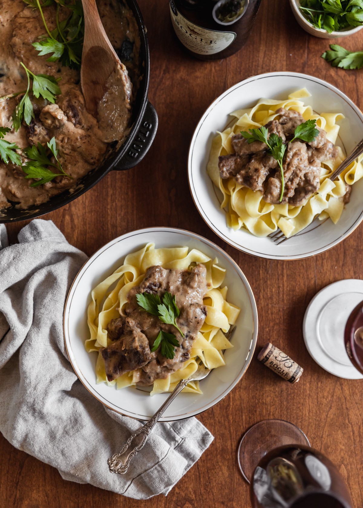 An overhead image of two white bowls of egg noddles topped with beef stroganoff with shallots and brandy on a wood table. In the upper left corner is a skillet with more stroganoff. Next to the bowls is a couple glasses of wine, a beige linen, a cork, and a white bowl of parsley.
