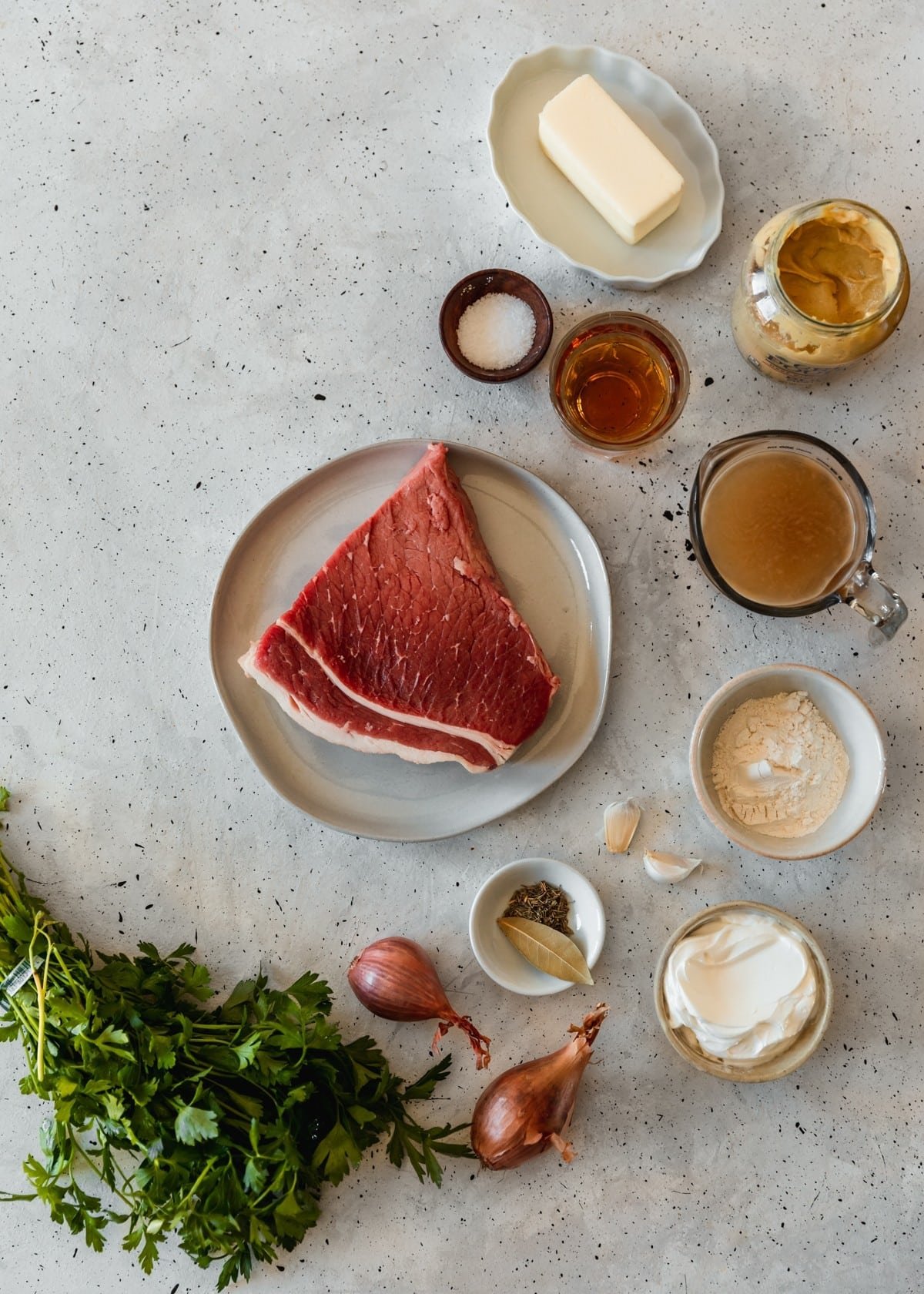 An overhead image of a white plate with a cut of steak on a grey table next to white bowls with flour and sour cream, a bunch of parsley, shallots, a measuring cup of beef broth, a glass of brandy, and a jar of Dijon mustard.