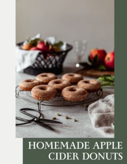 A side image of a circular cooling rack topped with baked apple cider donuts on a white speckled table with a wood tray of apples, black bowl of apples, and beige linen in the background.