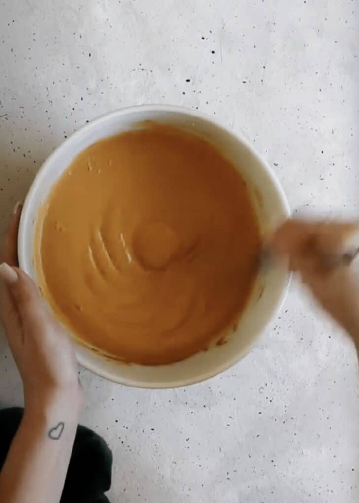A hand using a whisk to mix pumpkin filling in a white bowl on a grey counter.
