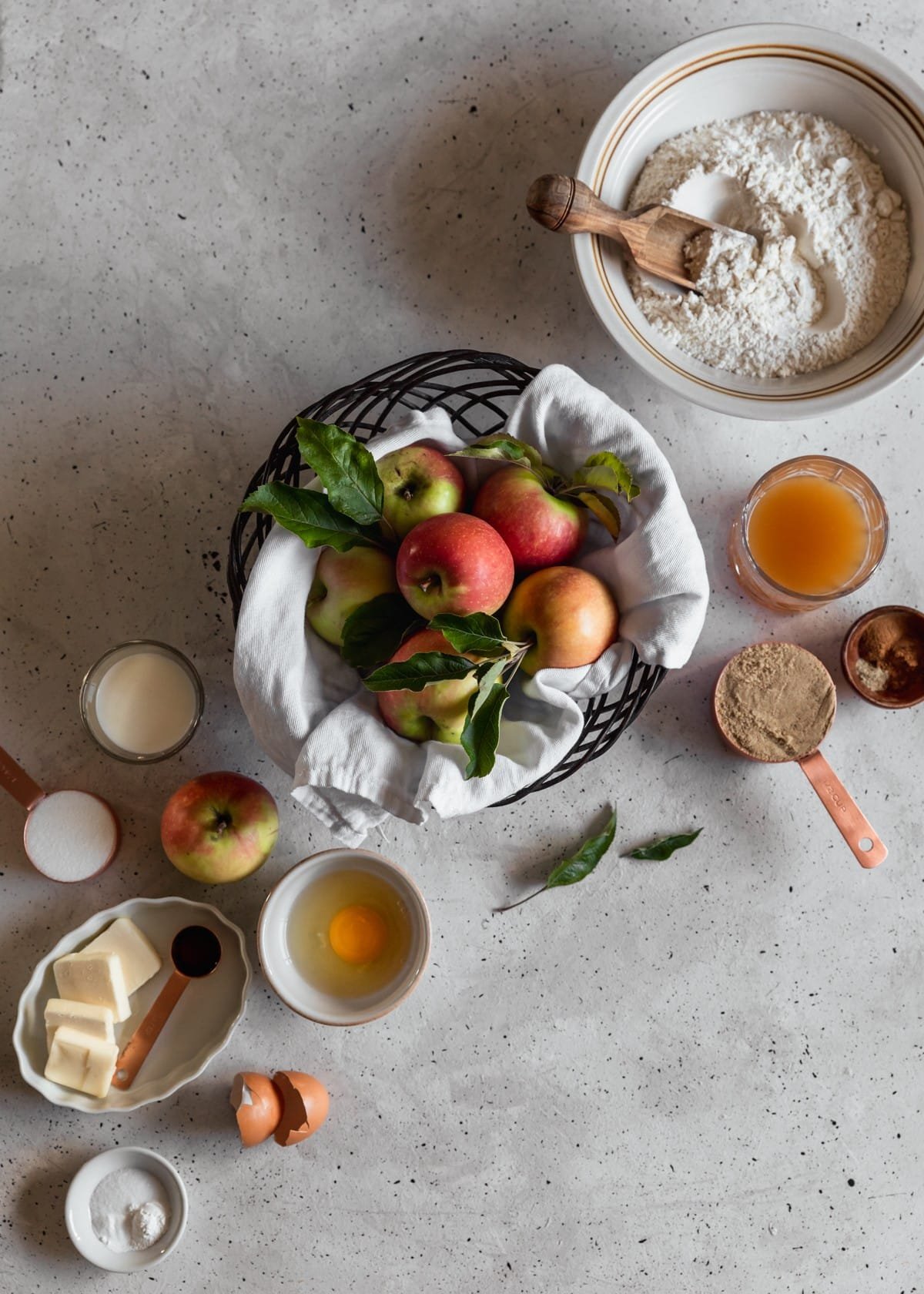 An overhead image of a black bowl of apples on a grey speckled table next to a glass of apple cider, copper measuring cups of sugar, and various white and grey bowls filled with baking ingredients.