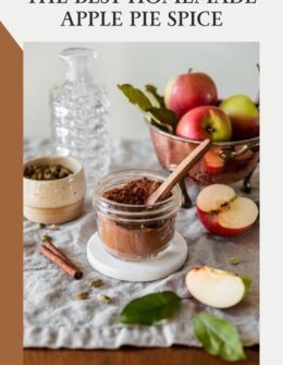 A side image of a jar of homemade apple pie spice on a beige linen next to a copper colander of apples, sliced apples, a white bowl of cardamom pods, and a glass vase.