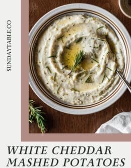 White cheddar mashed potatoes with butter and rosemary in a white bowl on a wood table.