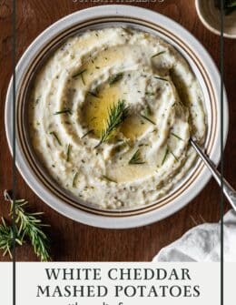 White cheddar mashed potatoes with butter and rosemary in a white bowl on a wood table.