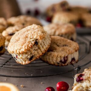 A very closeup image of a cranberry orange scone leaning on another scone on a vintage cooling rack. The rack is on a beige linen with more scones in the background and cranberries and an orange in the front.