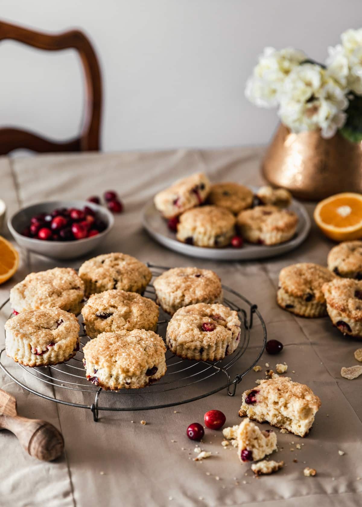 A vintage metal cooling rack topped with berry and citrus biscuits on a beige linen with a place of biscuits, various biscuits, a gold pot of white hydrangeas, and a wood spoon surrounding the rack.