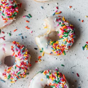 A closeup overhead image of funfetti donuts on a light grey counter. One of the donuts has a bite out of it.
