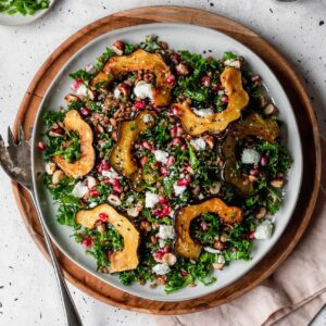 A very closeup image of a white plate with farro salad with kale, acorn squash, and pomegranates on a wood tray placed on a grey table next to a light pink linen.