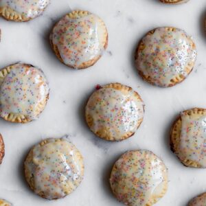 A very closeup image of rows of strawberry balsamic hand pies with white glaze and rainbow sprinkles on a white marble table.