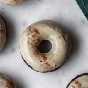 A very closeup image of a chocolate donut with brown glaze and a sprinkle of spices on a white marble counter with more donuts off to the side.