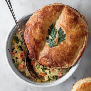 A very closeup image of a white bowl of chicken pot pie topped with puff pastry on a marble counter.