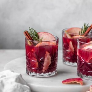 A side image of a glass of red wine sangria garnished with cinnamon and rosemary on a marble counter next to a white linen and two more glasses off to the side.