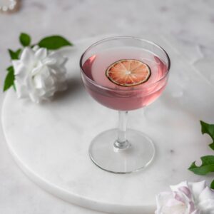 A side image of a prickly pear vodka sour in a coupe glass on a white marble counter next to white roses.