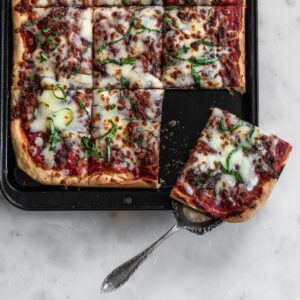 A closeup image of a sheet pan with Sicilian pizza on a marble counter. In the right corner, a pie server is holding up a slice of pizza.