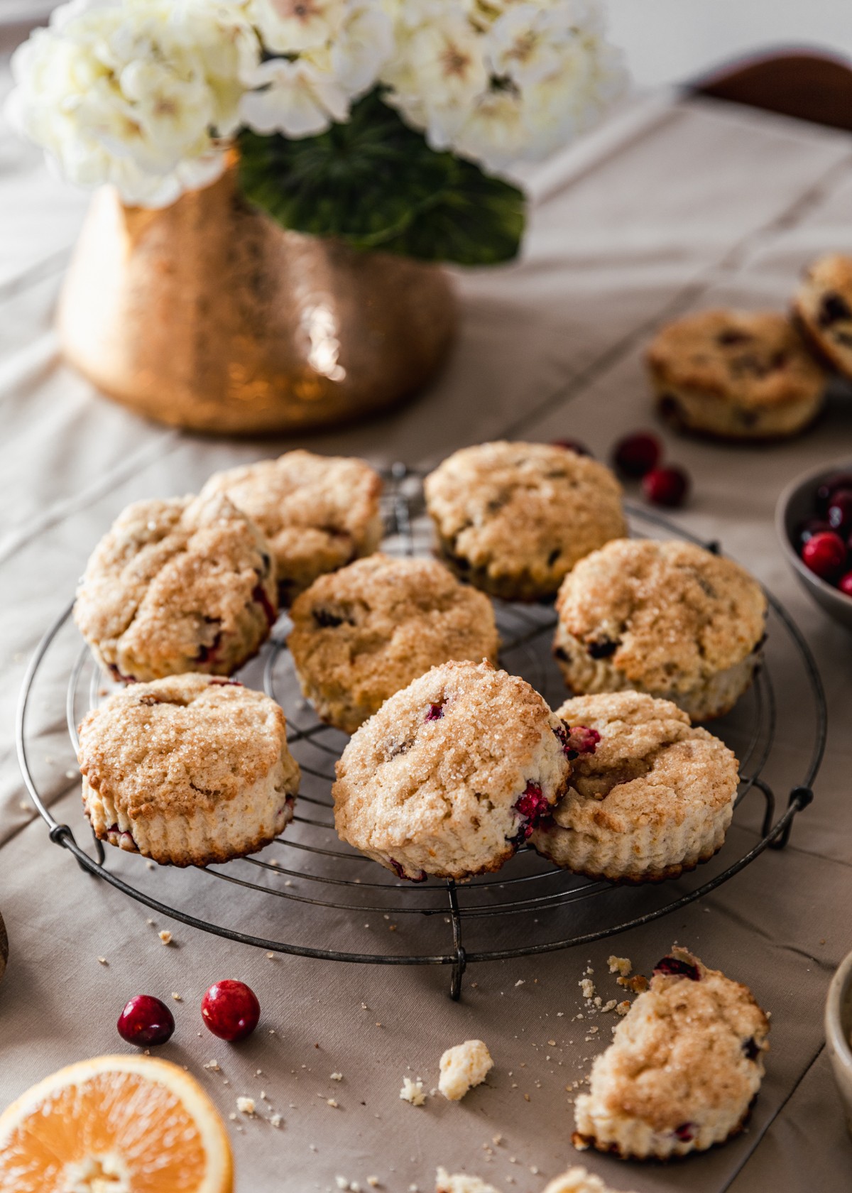 A side image of a metal circle cooling rack topped with cranberry orange scones on a beige table cloth next to a crumbled scone, half an orange, a bowl of cranberries, and a gold pot with white hydrangeas.