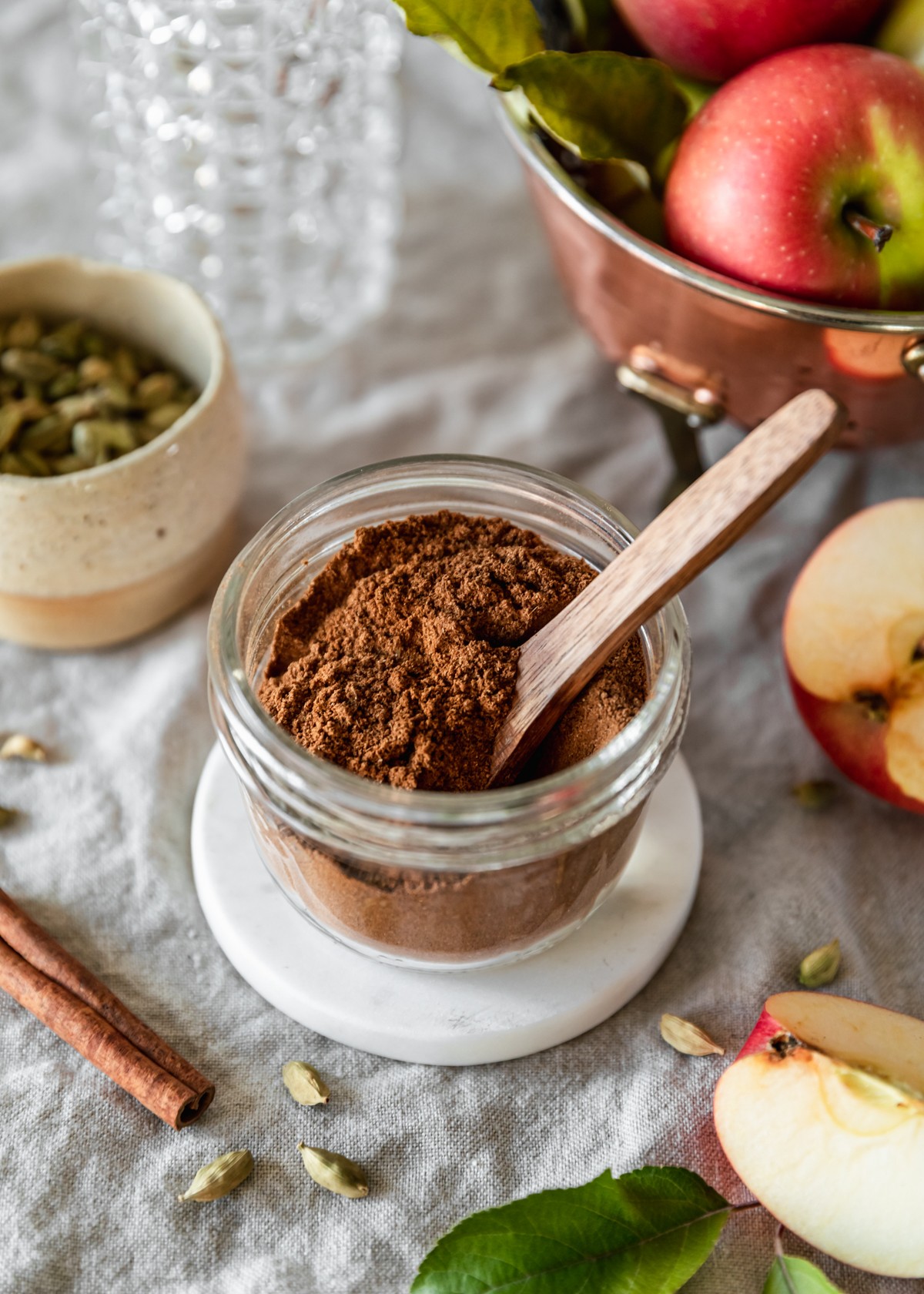 A side image of a jar of homemade apple pie spice on a beige linen next to a copper colander, sliced apple, cream bowl of cardamom pods, and a cinnamon stick.