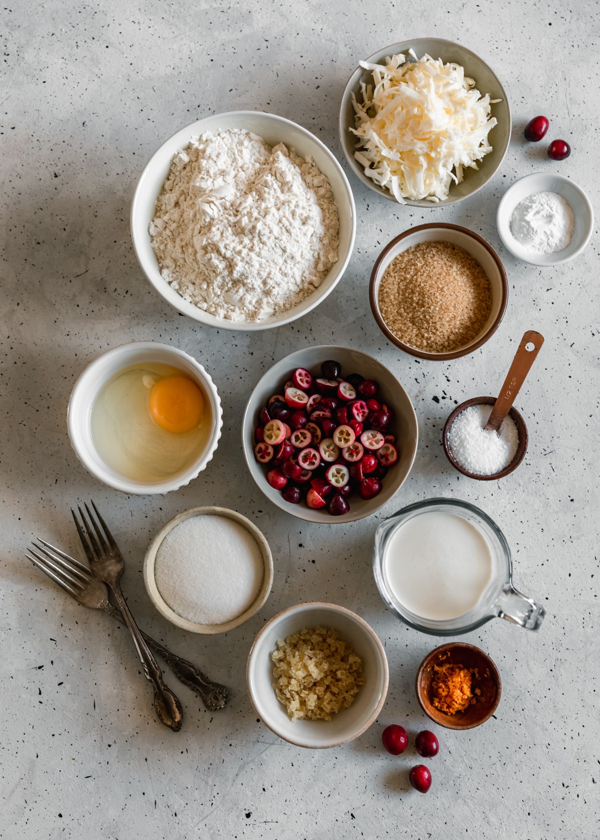 An overhead image of various white and grey bowls with baking ingredients, a grey bowl of cranberries, two vintage forks, and a wood bowl of citrus zest on a grey table.