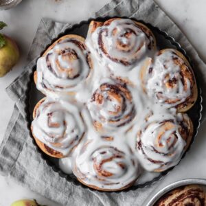 A closeup overhead image of a black circular tin of apple butter cinnamon rolls with frosting on a beige linen with a marble background.