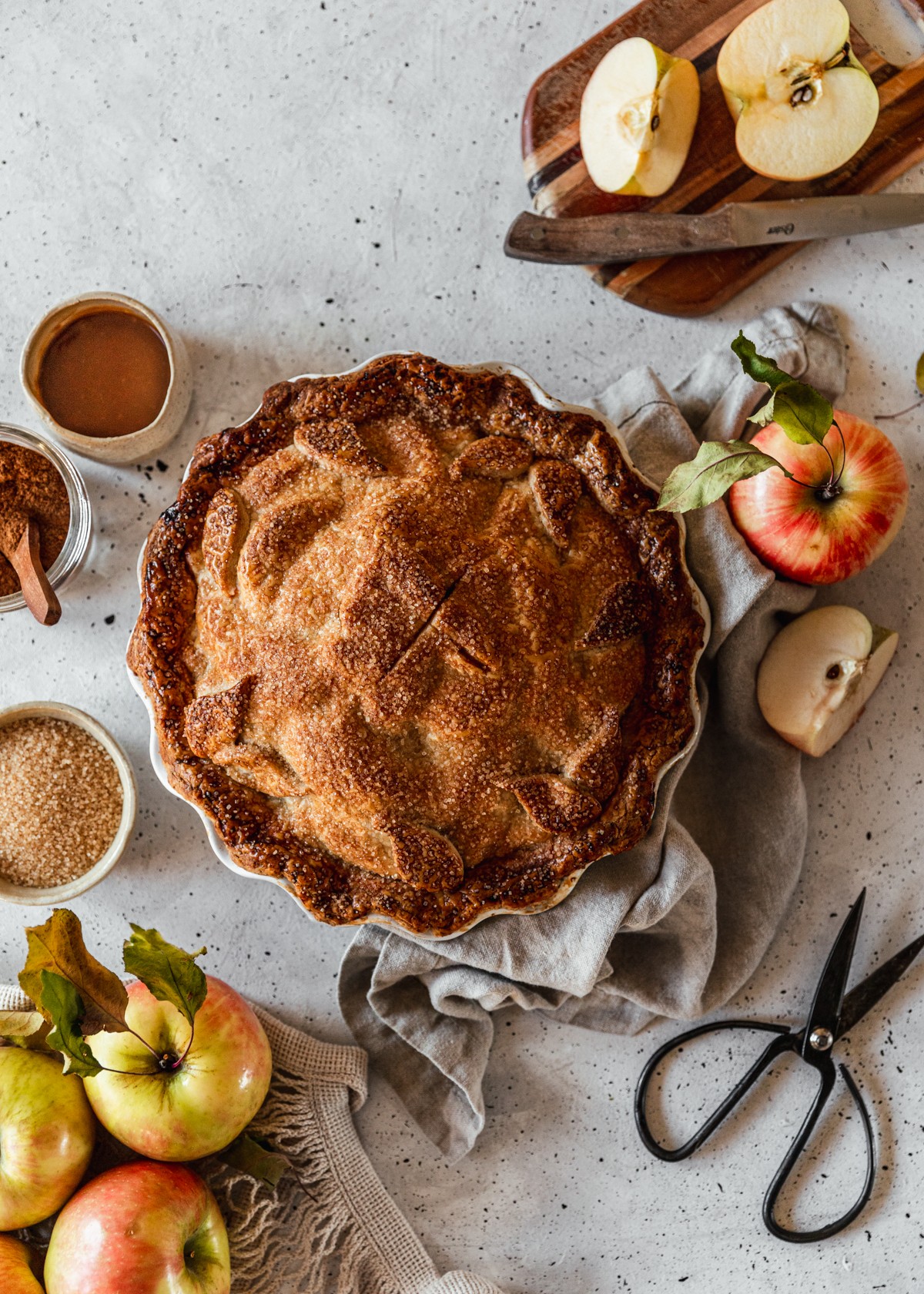 An overhead image of caramel apple pie in a white dish on a white speckled table next to a cutting board with a sliced apple, a few Honeycrisp apples, a bowl of caramel, a jar of apple pie spice, a pair of vintage black scissors, and a beige linen.
