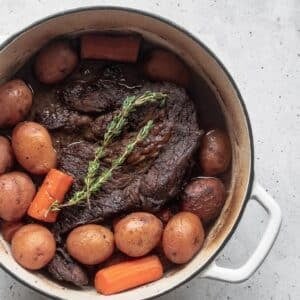 A closeup overhead image of half of a white pot filled with pot roast, potatoes, carrots, and thyme on a white speckled table.