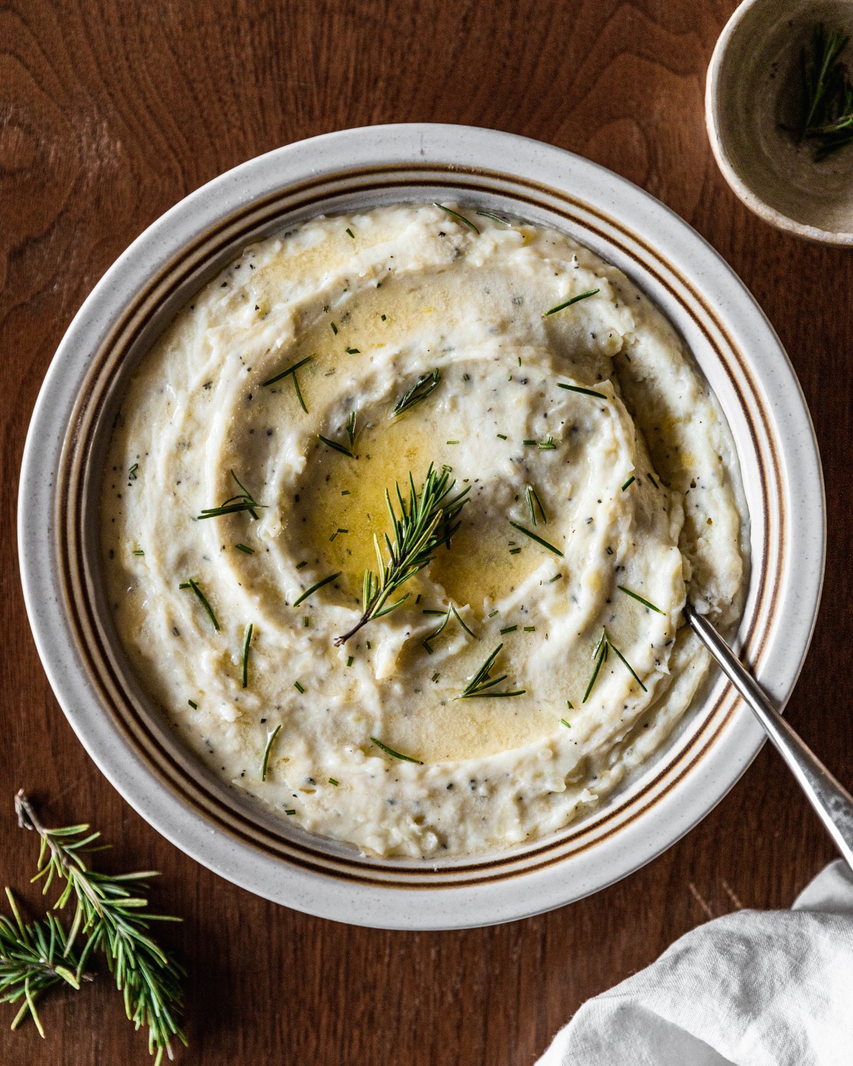 A very closeup overhead image of a white bowl of cheddar mashed potatoes garnished with butter and rosemary on a wood table next to a white linen and sprigs of rosemary.