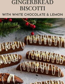 A closeup image of rows of gingerbread biscotti drizzled with white chocolate and lemon on a white speckled table with a pine branch in the background.