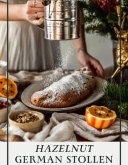A woman dusting powdered sugar over hazelnut German stollen next to greenery, oranges, and beige linens.