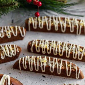 A closeup of a row of gingerbread biscotti on a white table with pine needles and red berries in the background.