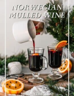 A woman pouring Norwegian mulled wine into a glass on a wood board next to oranges, greenery, and a beige napkin.