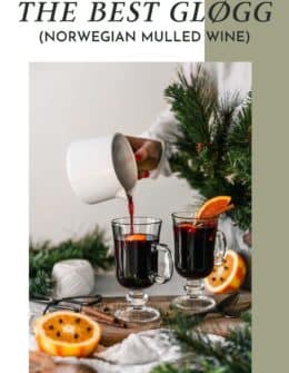 A woman pouring Norwegian mulled wine into a glass on a wood board next to oranges, greenery, and a beige napkin.