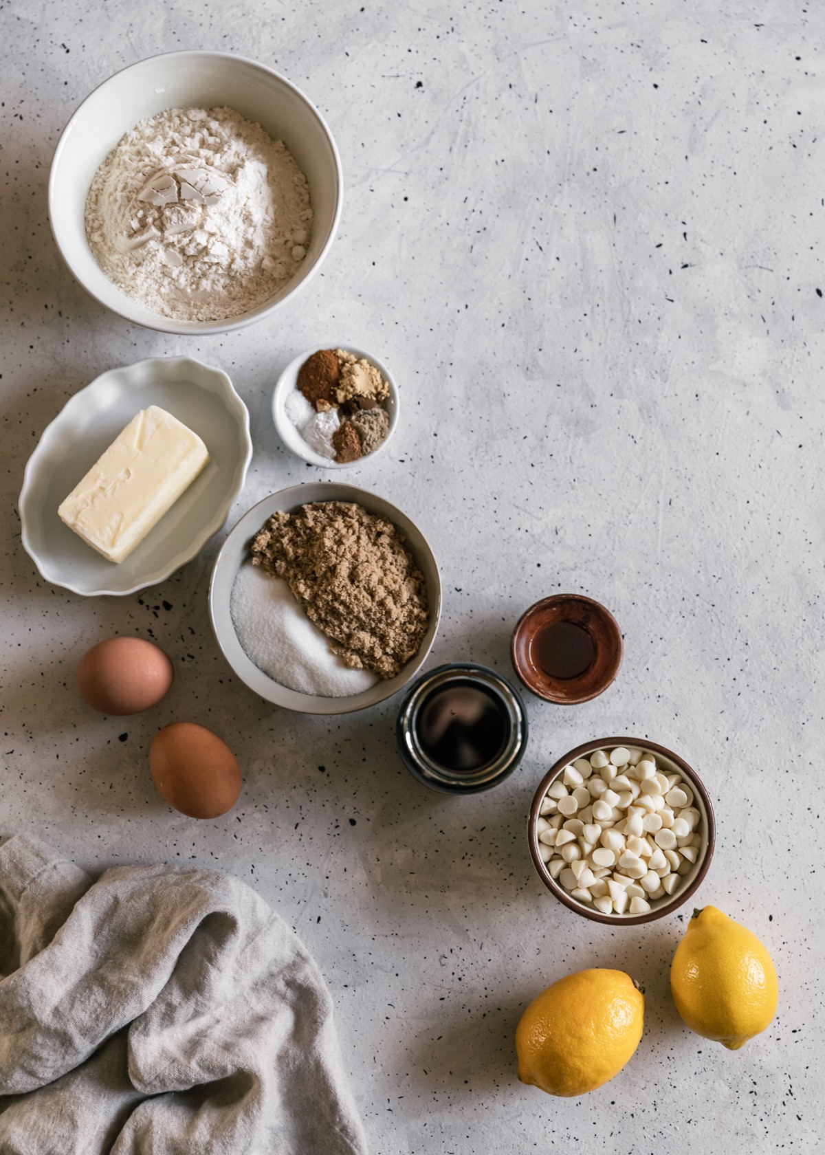 Various white bowls filled with flour, white sugar, brown sugar, spices, and butter on a grey speckled counter next to a jar of molasses, wood bowl of vanilla, eggs, 2 lemons, and a brown bowl of white chocolate.