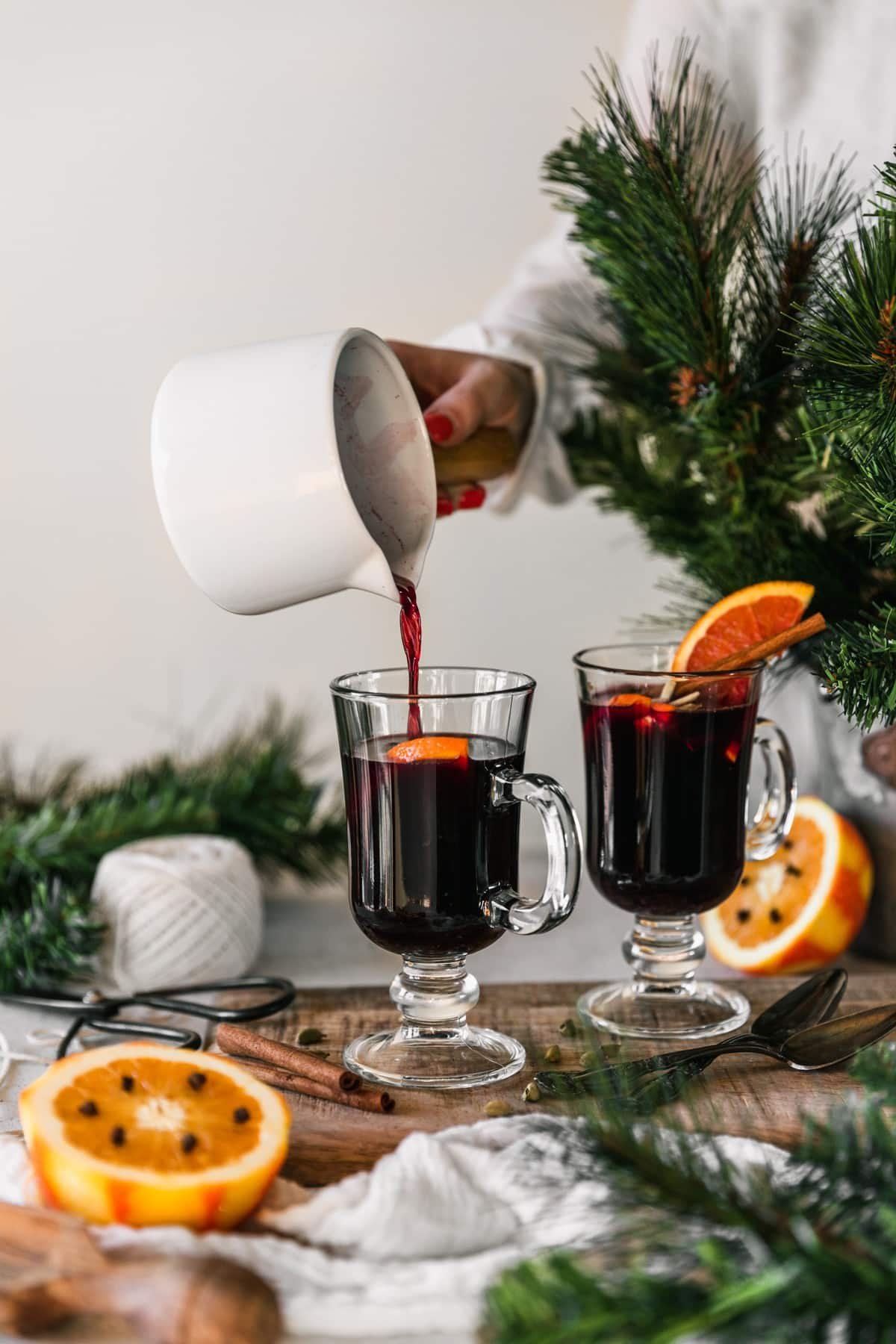 A side image of a woman's hand pouring gløgg from a white pot into a mug on a wood board next to another mug of gløgg, pine branches, halved oranges, and a roll of white twine with a white wall in the background.