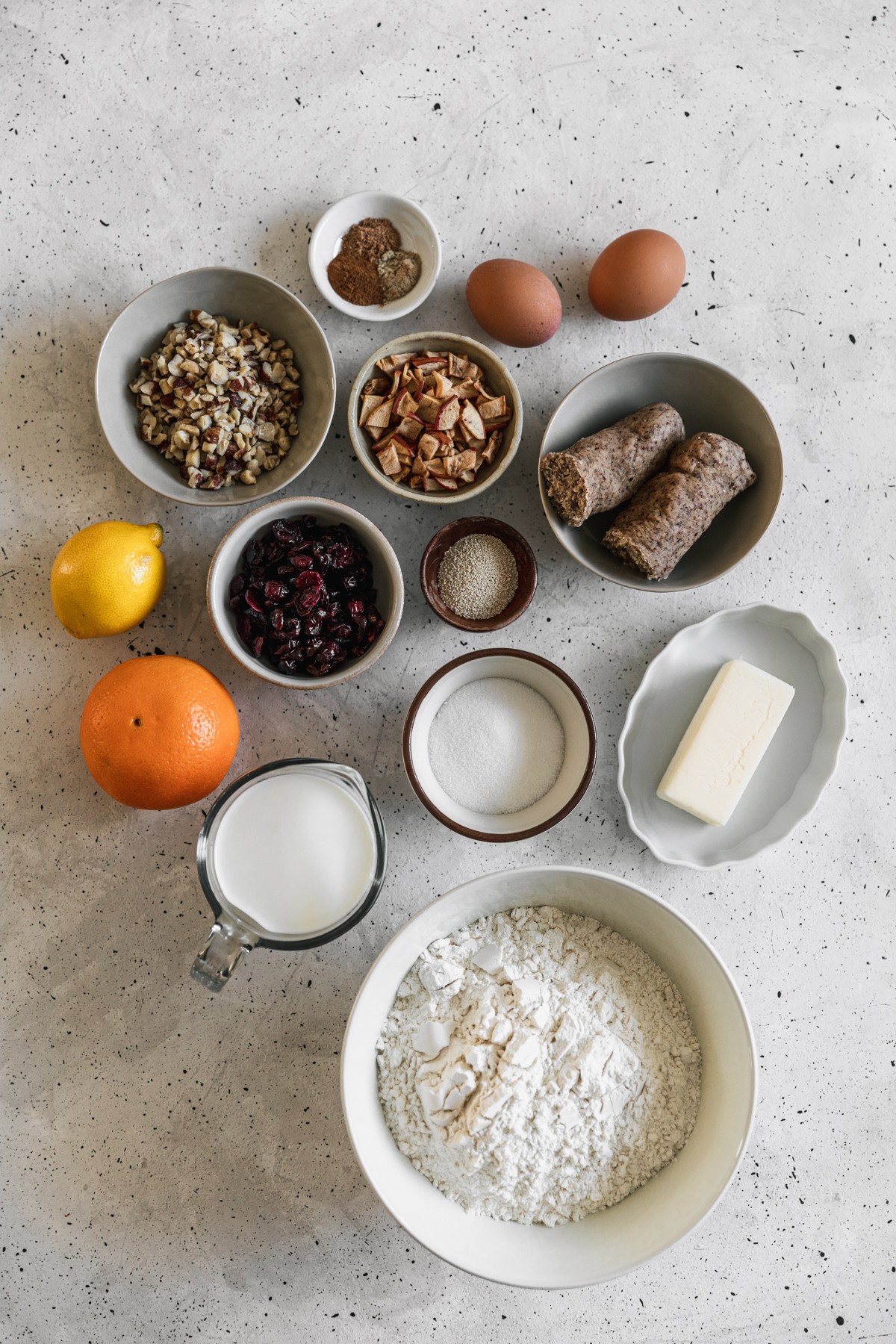 An overhead image of various white bowls with flour, dried apples, dried cranberries, butter, sugar, and spices on a speckled white table next to a measuring cup of milk, an orange, a lemon, and a wood bowl of yeast.
