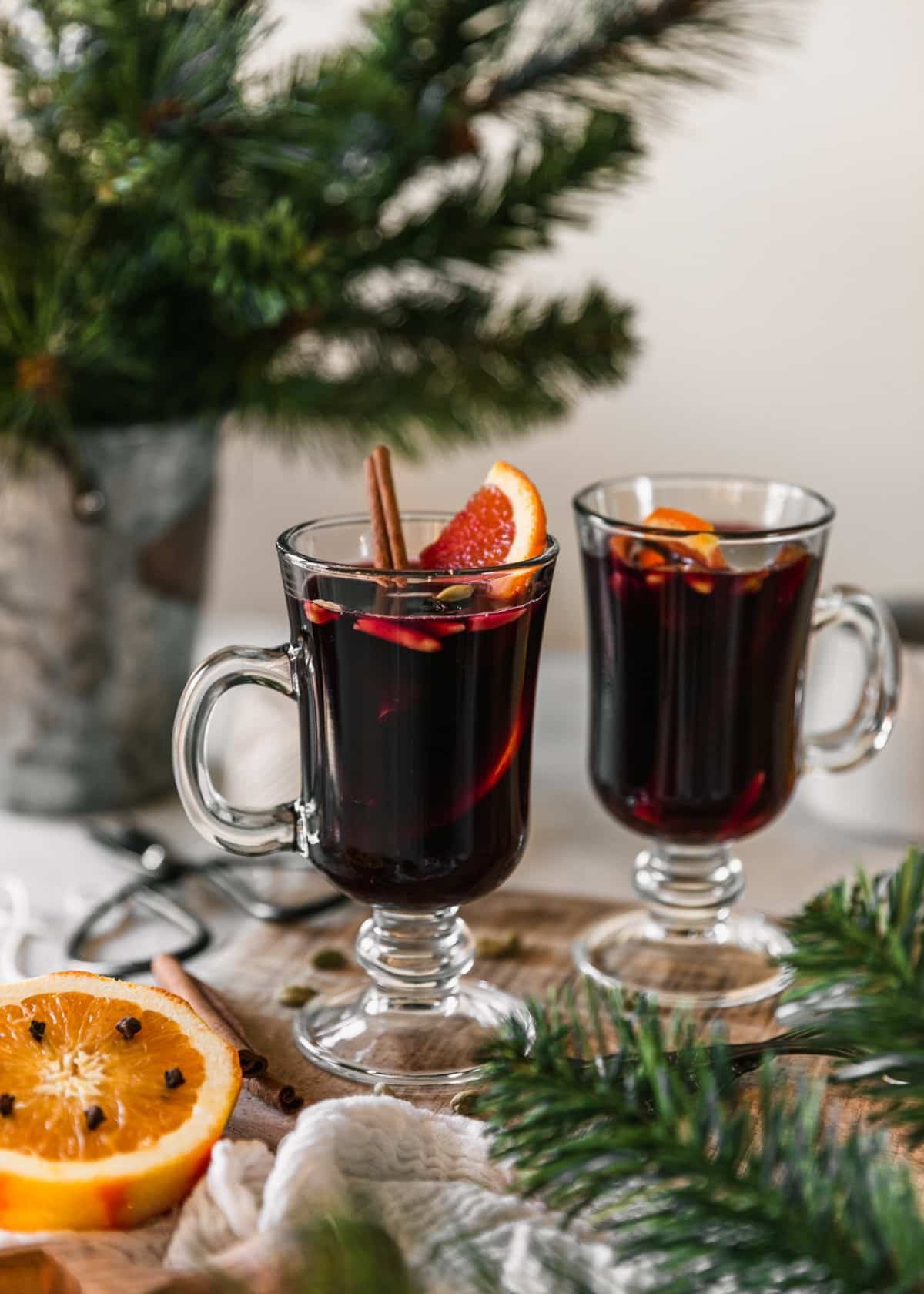 A side image of two glasses of gløgg on a wood board on a white counter next to a metal pot with pine branches, a halved orange, a pair of scissors, and a white linen.