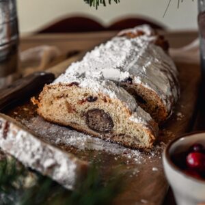 A closeup image of sliced stollen on a wood board next to a white bowl of cranberries and a pine branch.