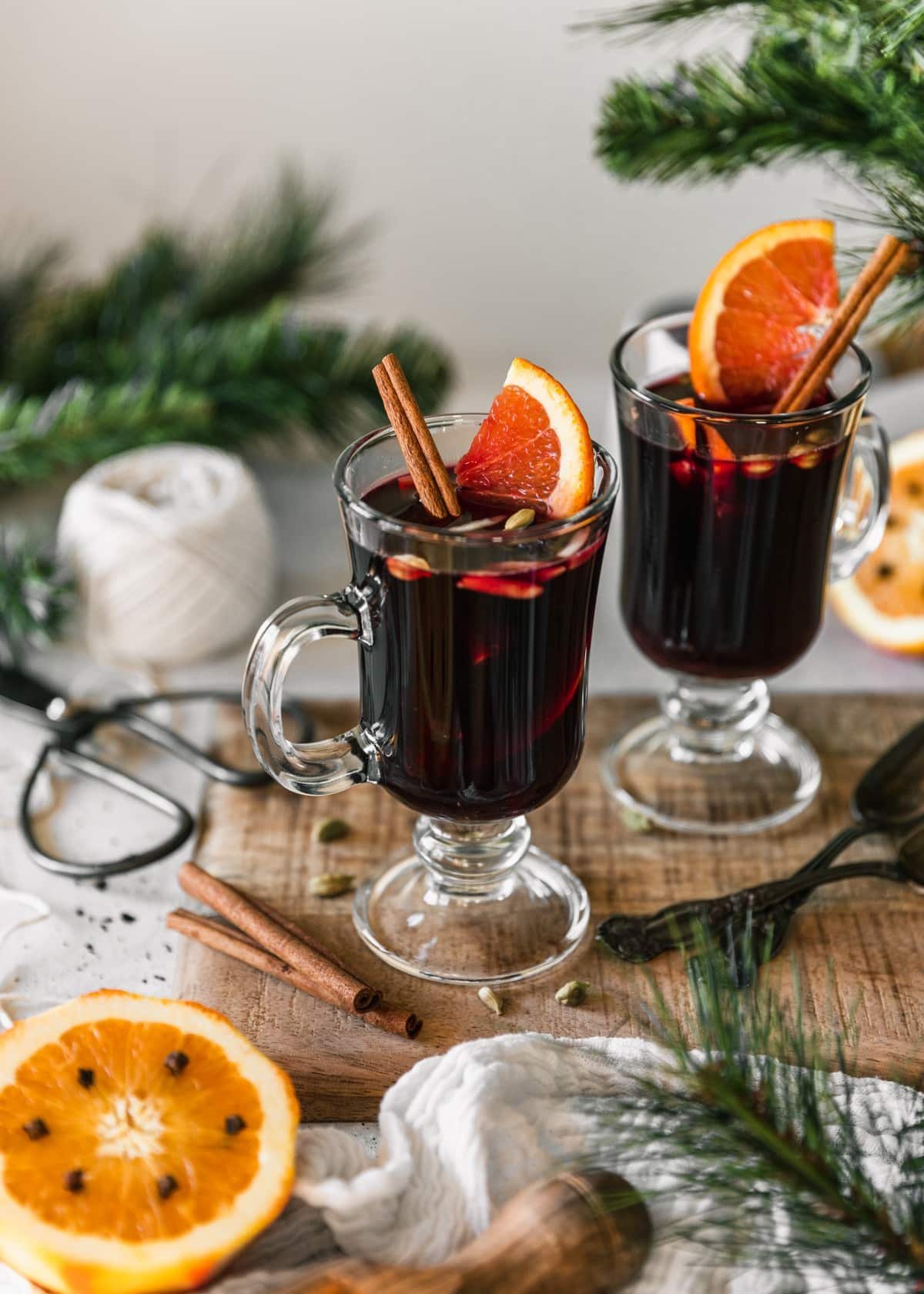 A side image of two glasses of gløgg garnished with cinnamon sticks and orange slices on a wood board with a white background next to pine branches, halved oranges, a white roll of twine, and a white linen.