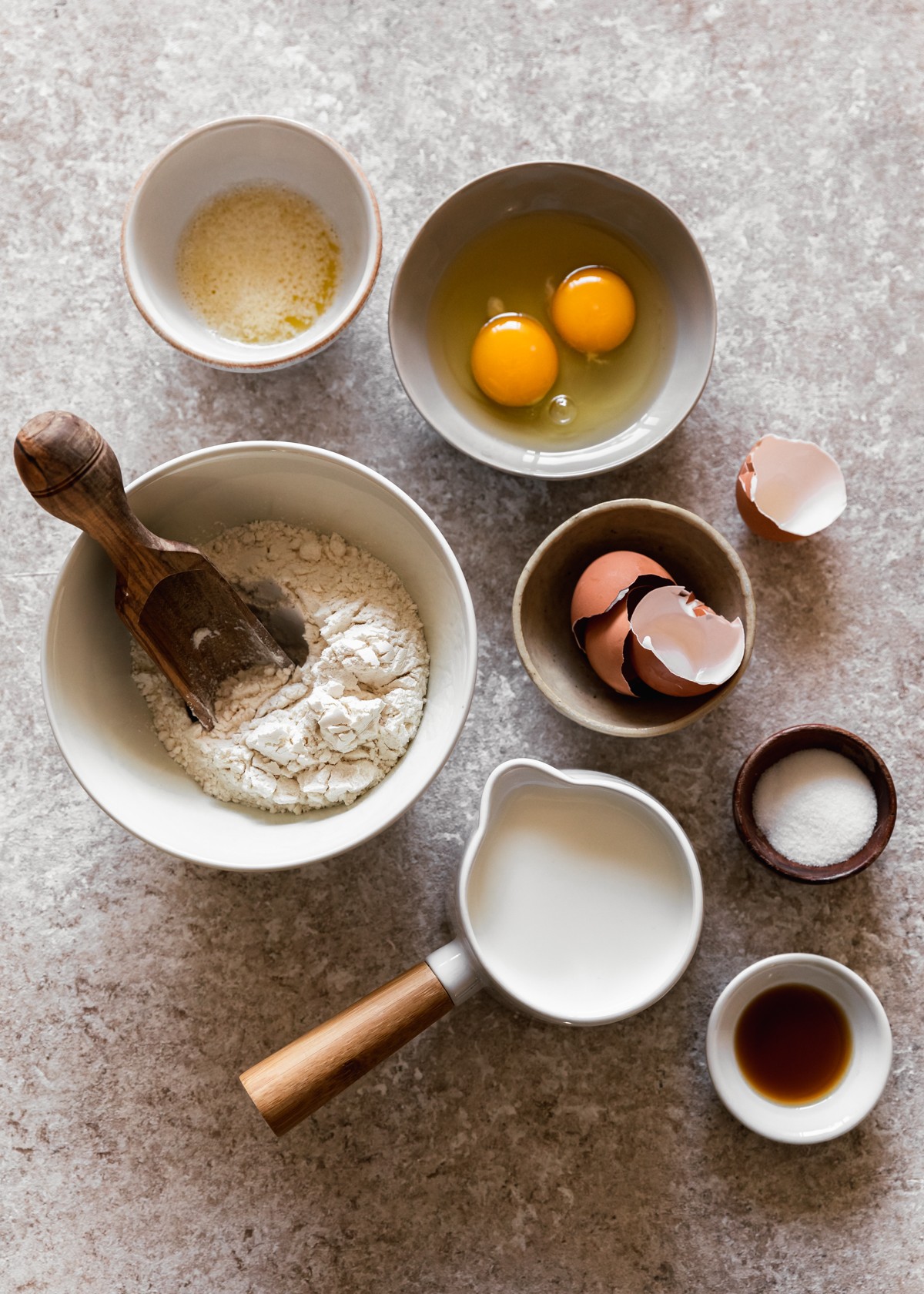 An overhead image of a white bowl of flour with a wood scoop, a cream bowl of egg shells, a white cup of milk, a wood bowl of salt, a beige bowl of eggs, and a white bowl of melted butter on a beige counter.