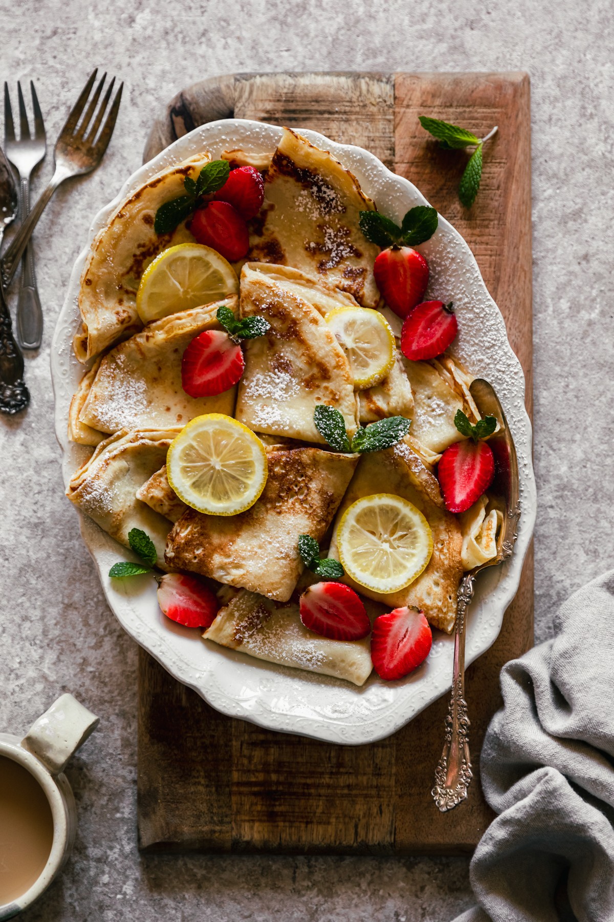 An overhead image of a white oval plate of Norwegian pancakes with berries, lemon slices, and mint on a wood board with a speckled beige background next to a beige napkin and vintage forks.