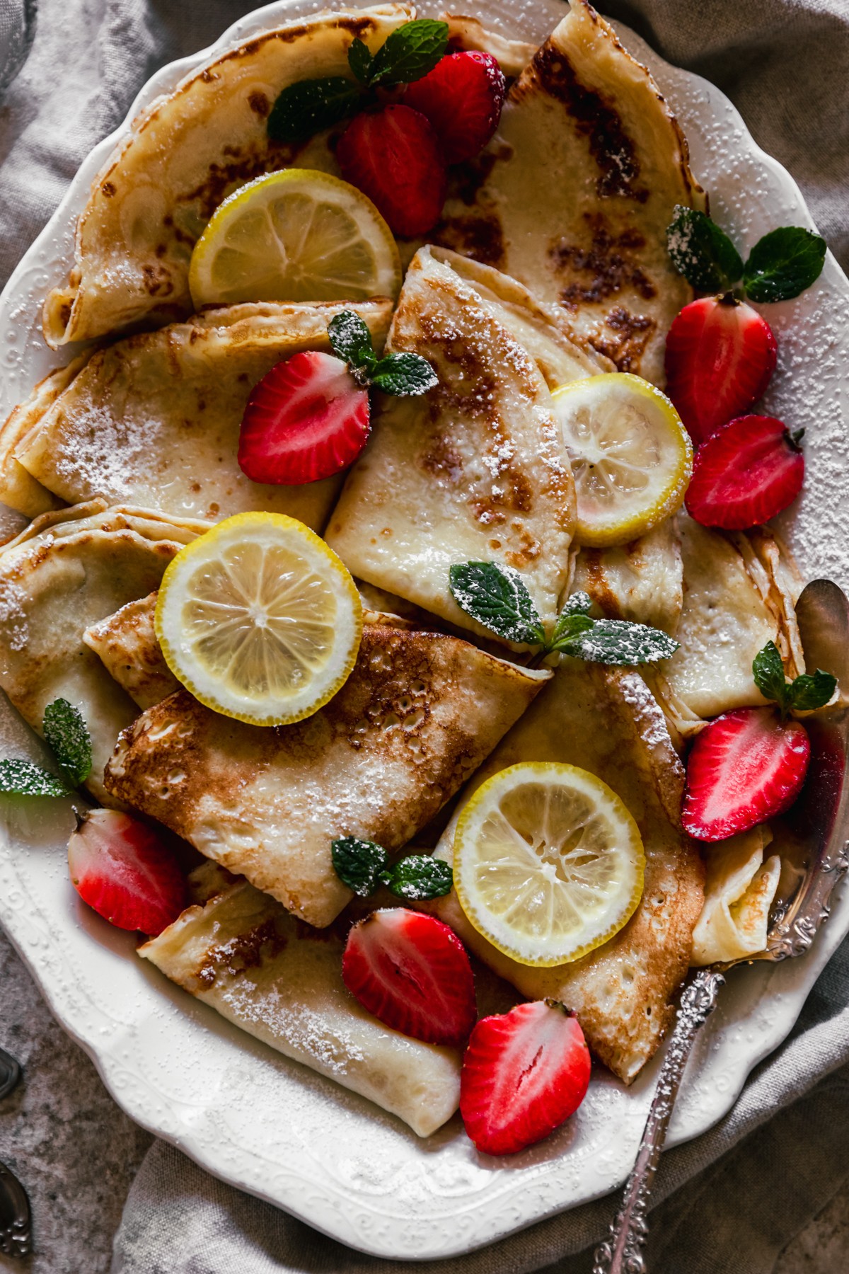 A very closeup image of Norwegian pancakes with strawberries and lemon slices on a white oval plate with a vintage serving spoon in the right lower corner. The background is a beige linen and tan speckled table.