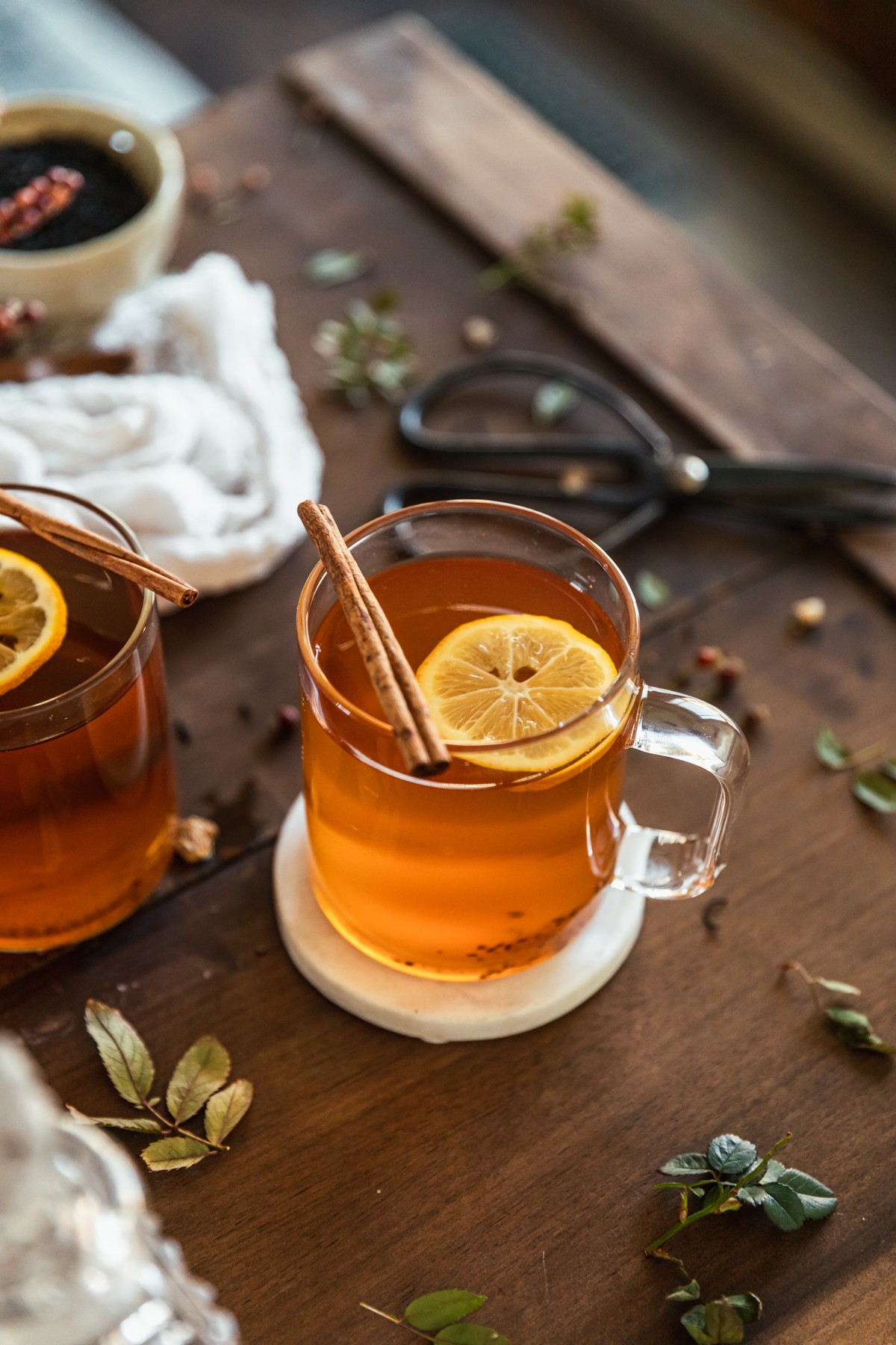 A close up 45-degree shot of a tea hot toddy with a lemon slice and a cinnamon stick on a wood table next to another toddy, a pair of vintage scissors, a white towel, and tea laves.