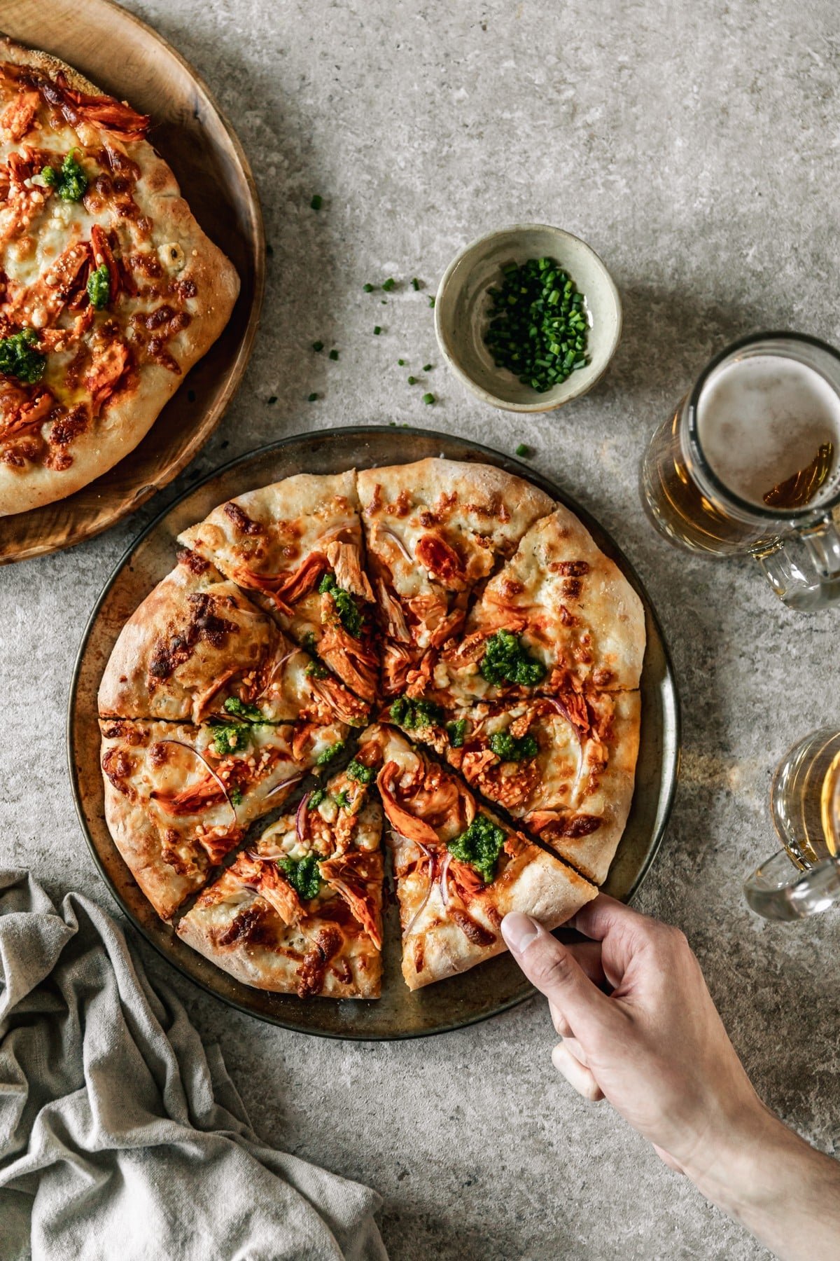 An overhead image of a man grabbing a slice of buffalo chicken pizza off of a metal tray on a beige counter next to a linen, mugs of beer, and another pizza.