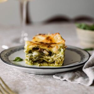 A closeup side image of a slice of pesto white lasagna on a white plate on a beige table next to a tan linen and white bowl of basil.