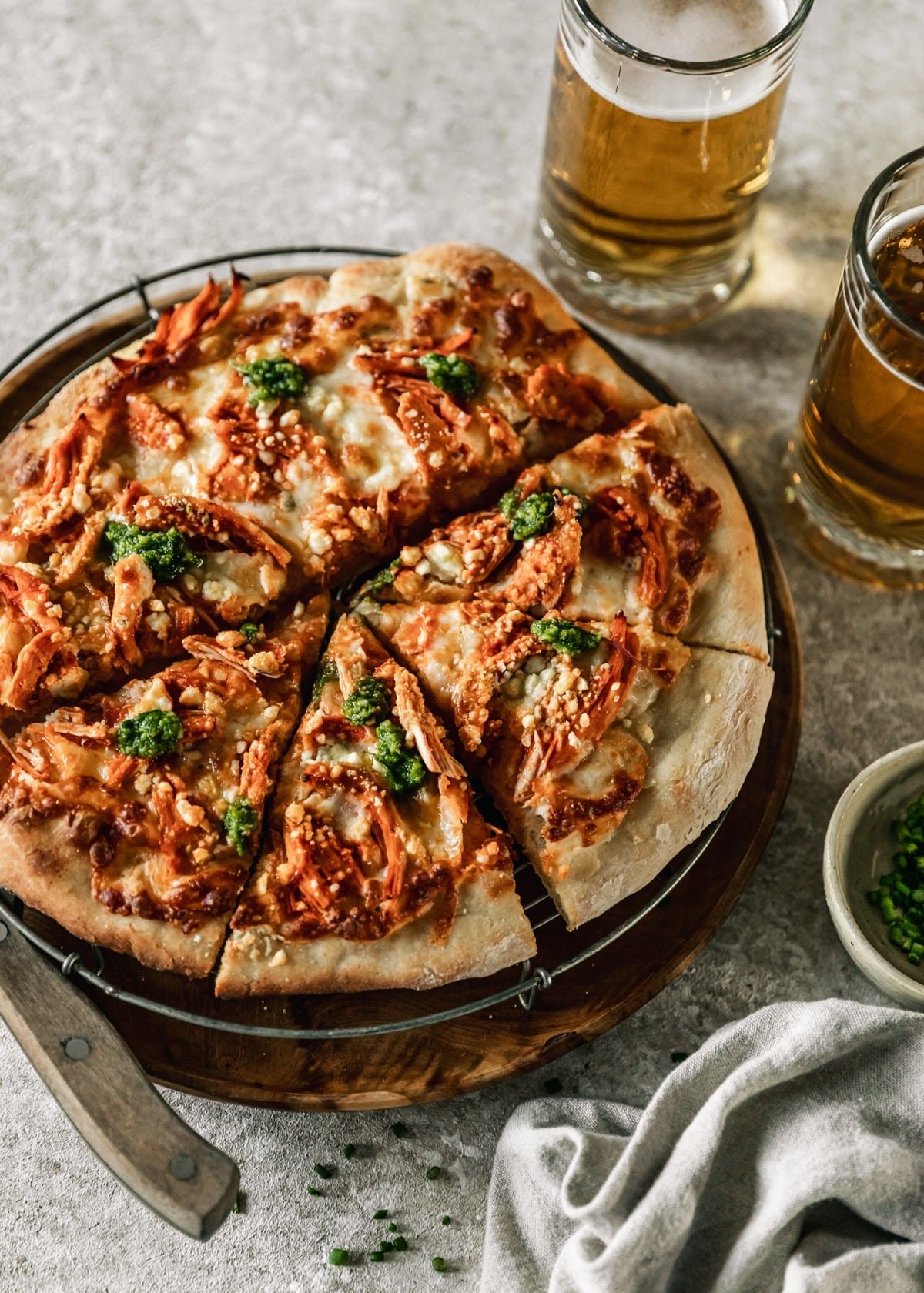 A side image of a buffalo chicken pizza with chive pesto on a wood and metal tray on a tan counter next to a white bowl of chives and two mugs of beer.