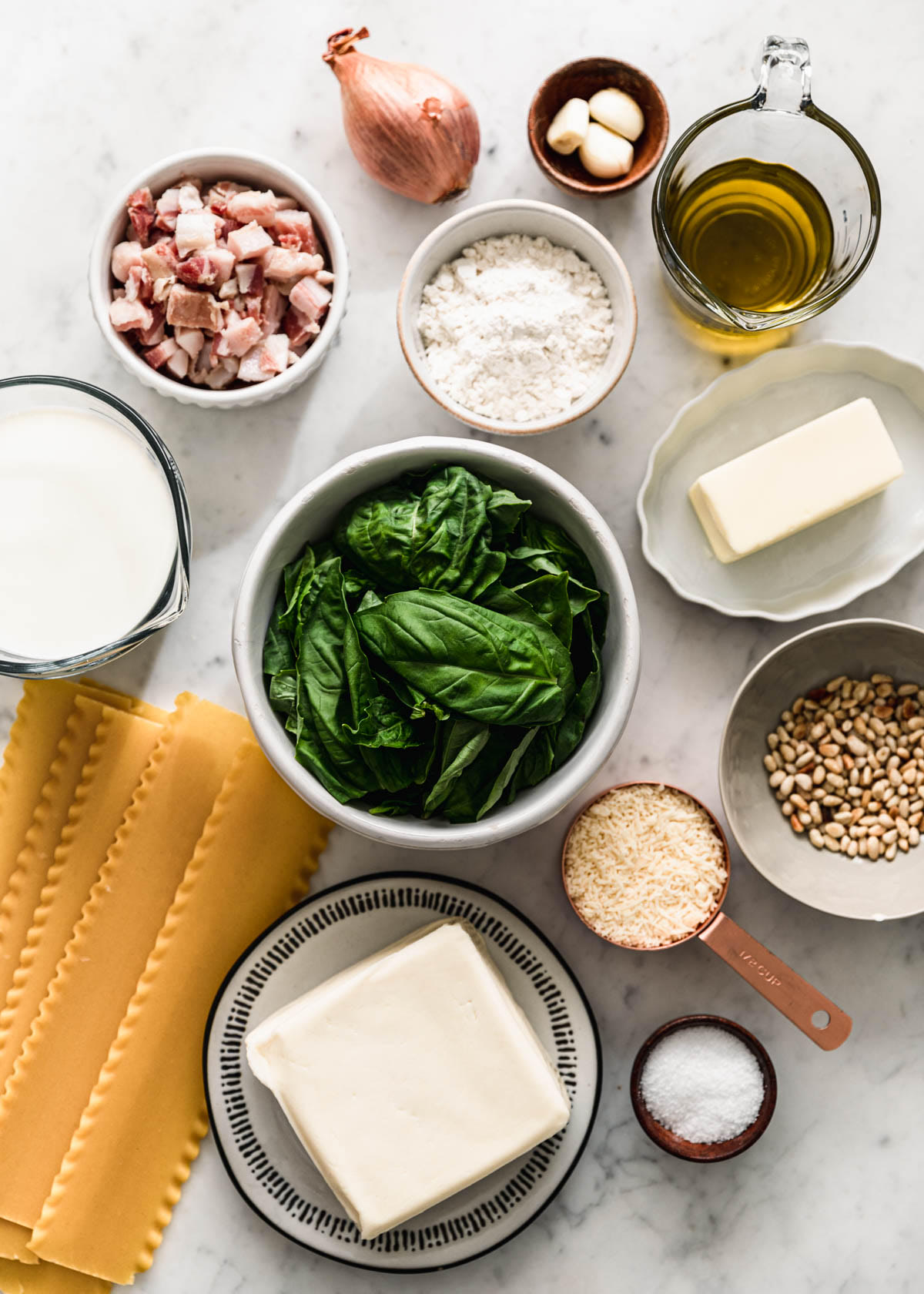 An overhead image of a cream-colored bowl of basil, a bowl of flour, a grey bowl of pine nuts, a measuring cup of Parmesan, measuring cups with milk and olive oil, a shallot, noodles, and a bowl of pancetta on a marble counter.