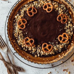 A closeup overhead image of a chocolate caramel tart with pretzels and peanuts on a white counter next to vintage forks.