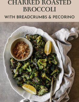 An overhead image of a white oval dish of roasted broccoli with lemon wedges and breadcrumbs on a brown table next to a tan napkin.